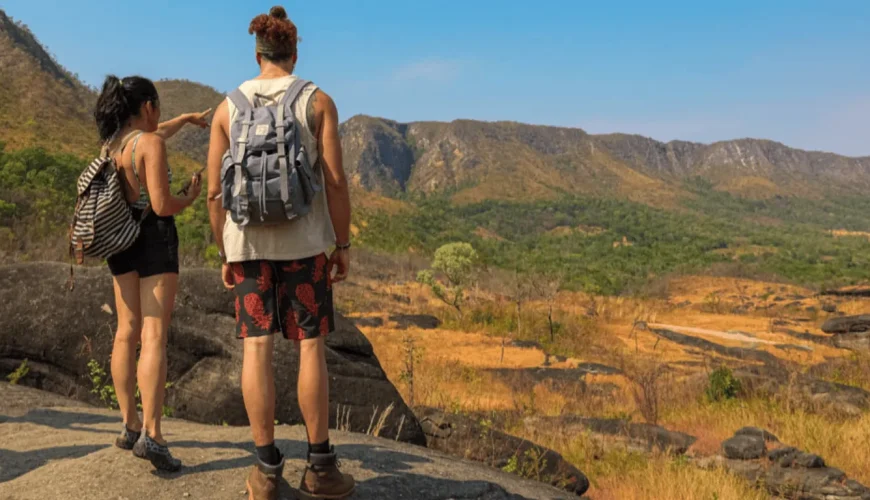 Dois viajantes de costas, com mochilas, observam a vasta paisagem do Cerrado a partir de um mirante rochoso na Chapada dos Veadeiros, com serras e vegetação nativa ao fundo sob céu azul.
