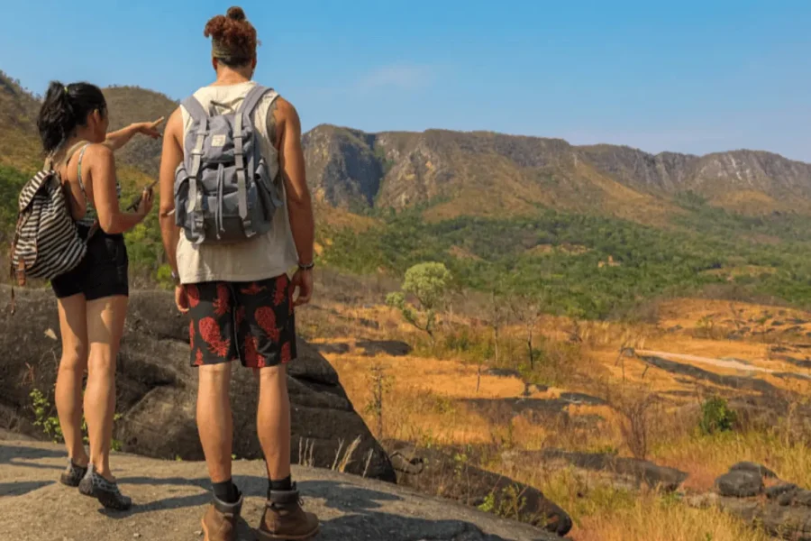 Dos viajeros de espaldas, con mochilas, observan el vasto paisaje del Cerrado desde un mirador rocoso en la Chapada dos Veadeiros, con sierras y vegetación nativa al fondo bajo un cielo azul.
