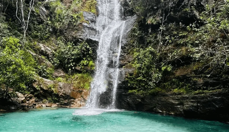 Cascata de água cristalina a despenhar-se sobre rochas entre vegetação nativa do Cerrado, formando uma piscina natural de águas turquesa na Chapada dos Veadeiros, Goiás.