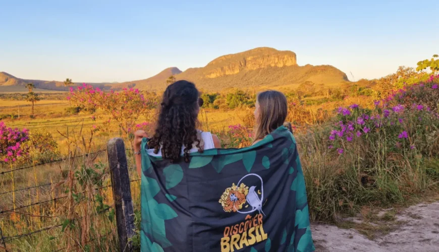 Duas viajantes de costas, partilhando uma bandeira da Discover Brasil, contemplam o pôr do sol dourado sobre as formações rochosas e as flores roxas do Cerrado na Chapada dos Veadeiros, Goiás.