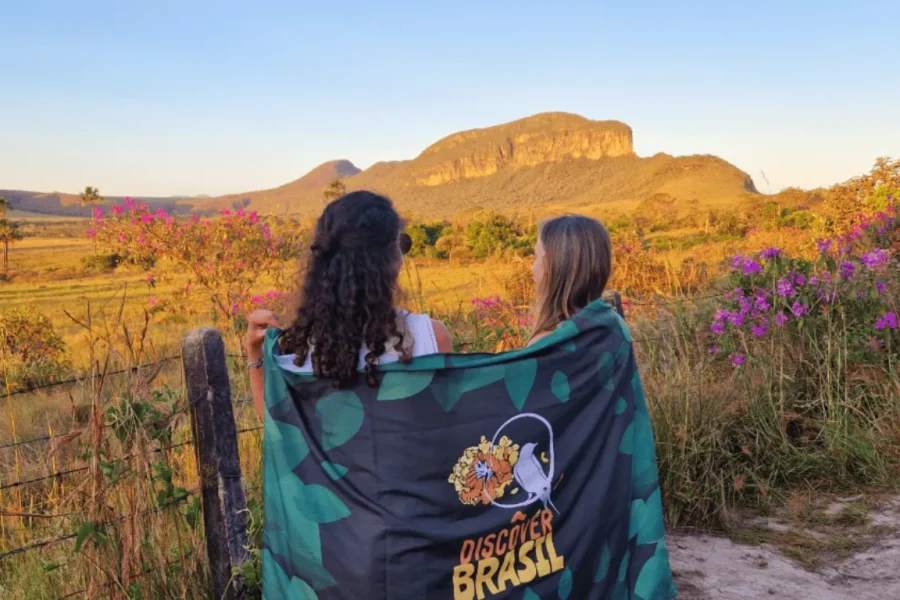 Dos viajeras de espaldas, compartiendo una bandera de Discover Brasil, contemplan el atardecer dorado sobre las formaciones rocosas y las flores silvestres moradas del Cerrado en la Chapada dos Veadeiros, Goiás.