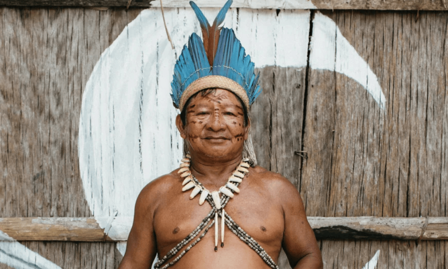Indigenous man from the Cipiá community in Amazonas, standing in front of a wooden wall, wearing a blue feather headdress and a traditional necklace, with facial body paint representing Amazonian culture.