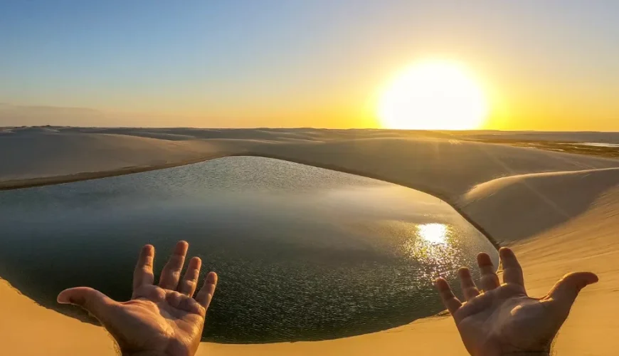 Mãos estendidas sobre uma lagoa cristalina nos Lençóis Maranhenses, com dunas brancas e pôr do sol dourado ao fundo