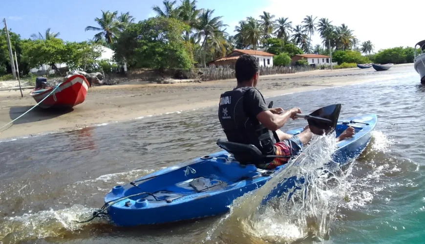 Homem a remar num caiaque azul no Rio Preguiças, com barcos e o Povoado Bar da Hora ao fundo.